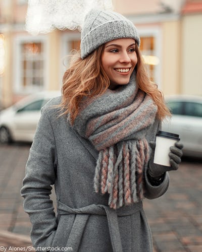Woman wearing a grey coat, with a chunky scarf around her neck, gloves, and a knitted beanie holding a cup of coffee