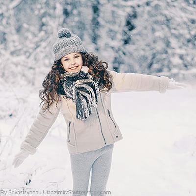 Young girl playing in the snow wearing a grey puffer coat, a knit scarf, gloves and a grey beanie with a pom pom on top