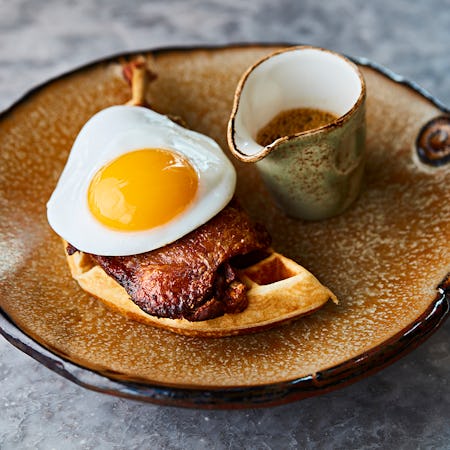 A brown plate serving up an enticing brunch: a fresh cooked egg over crisp bacon atop a half a waffle, with a ramekin of brown sauce